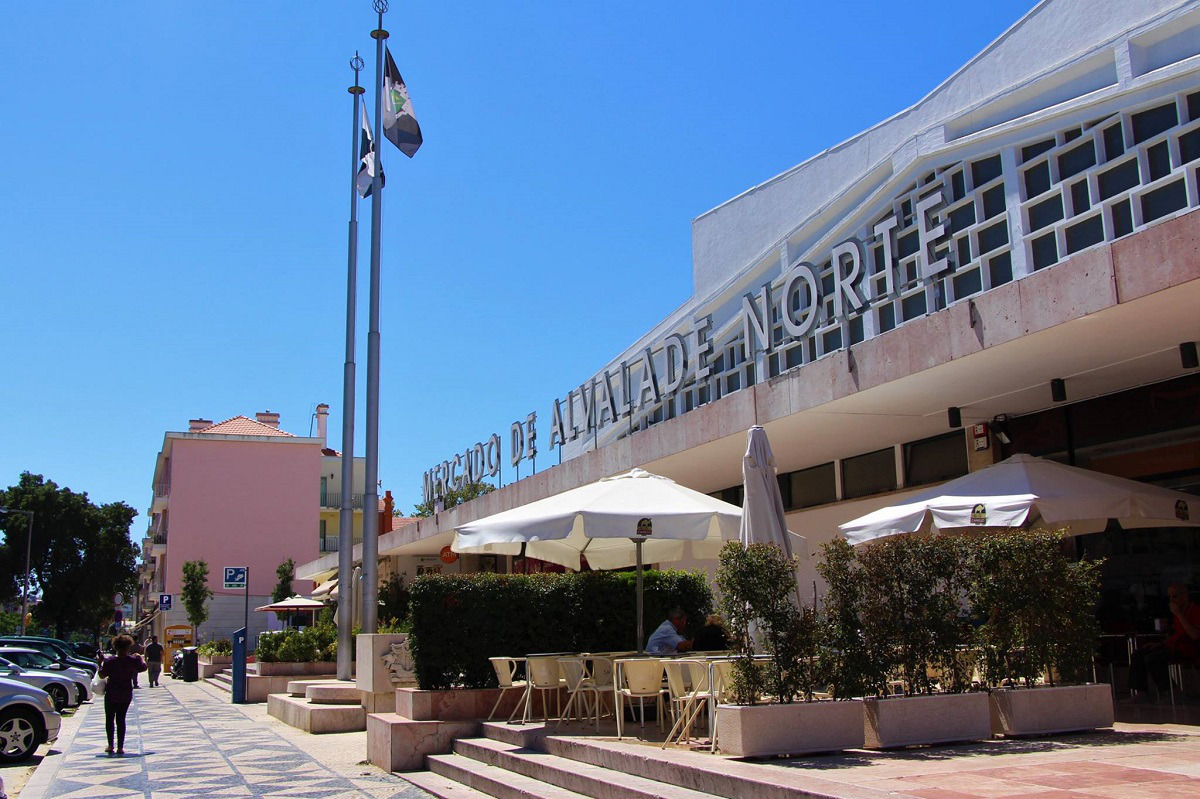 Fotografia da fachada do edifício, com a placa "Mercado de Alvalade Norte", área de esplanadas com guarda-sóis e mastros com bandeiras sob um céu azul claro