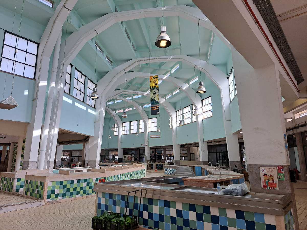 Interior do Mercado de Arroios com estrutura moderna, teto alto com arcos brancos, janelas grandes, iluminação ampla e área de alimentação com balcões de cores variadas.
