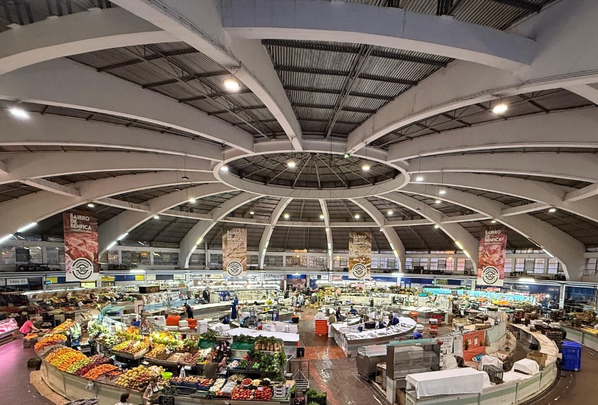 Imagem do Mercado de Benfica coberto, com várias bancas de frutas, legumes e outros produtos, sob uma estrutura de teto circular com iluminação moderna.