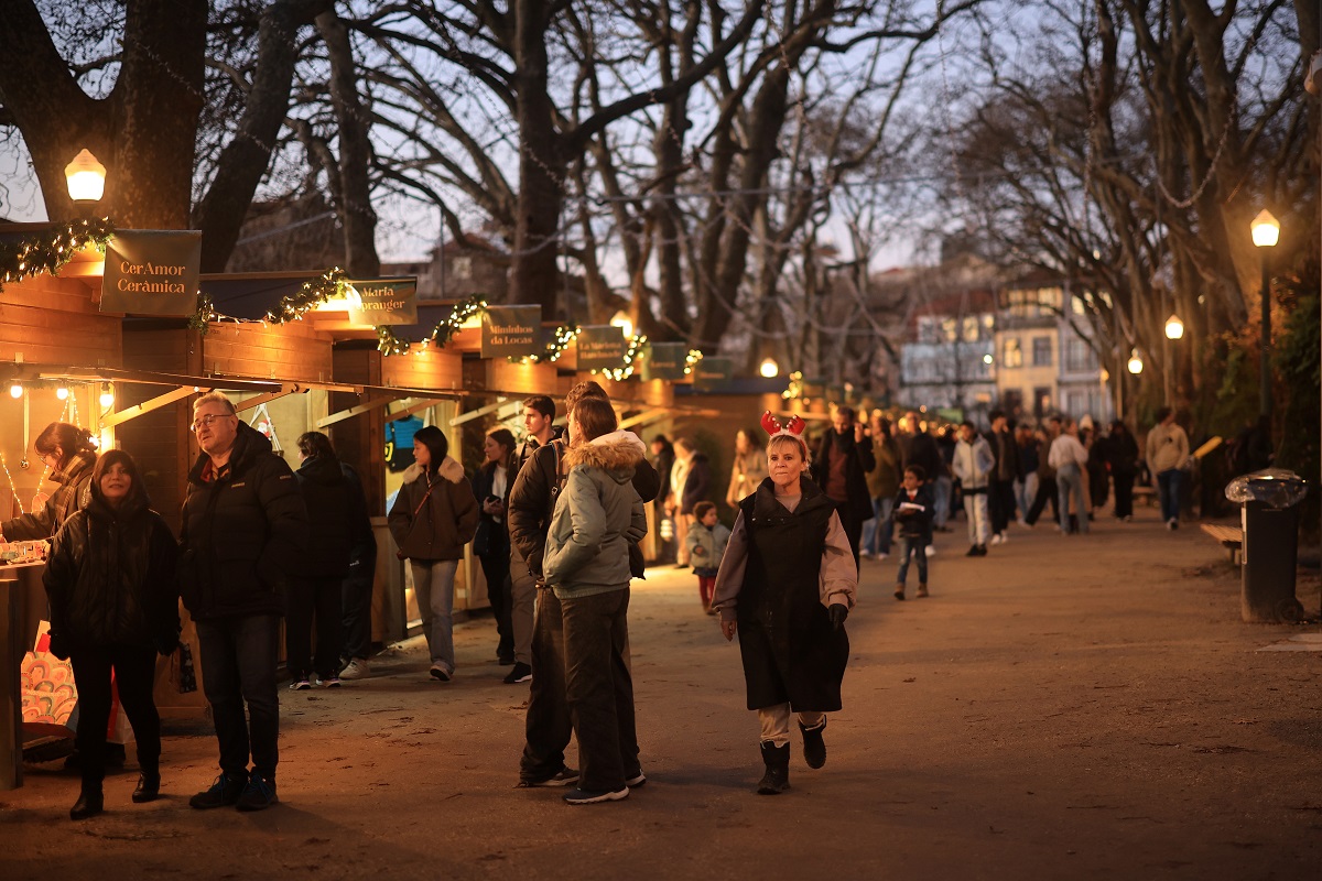 Imagem de um mercado de natal, num parque, à noite, com várias pessoas a passear e a aproveitar as tendas de comida e artesanato brilhando com luzes quentes, num ambiente acolhedor e festivo.