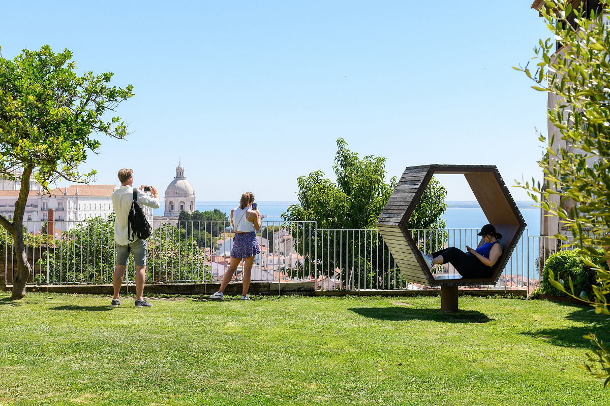 No Miradouro do Recolhimento, três pessoas aproveitando o espaço ao ar livre, com vista para o rio Tejo ao fundo. Enquanto duas delas tiram fotos, uma outra descansa numa cadeira em formato hexagonal.