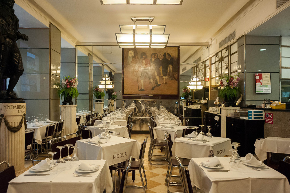 Interior elegante do Café Nicola, com mesas bem arrumadas, toalhas brancas e paredes espelhadas. Ao fundo da sala um grande quadro alusivo à vida do poeta Bocage. Arranjos florais realçam o ambiente clássico.