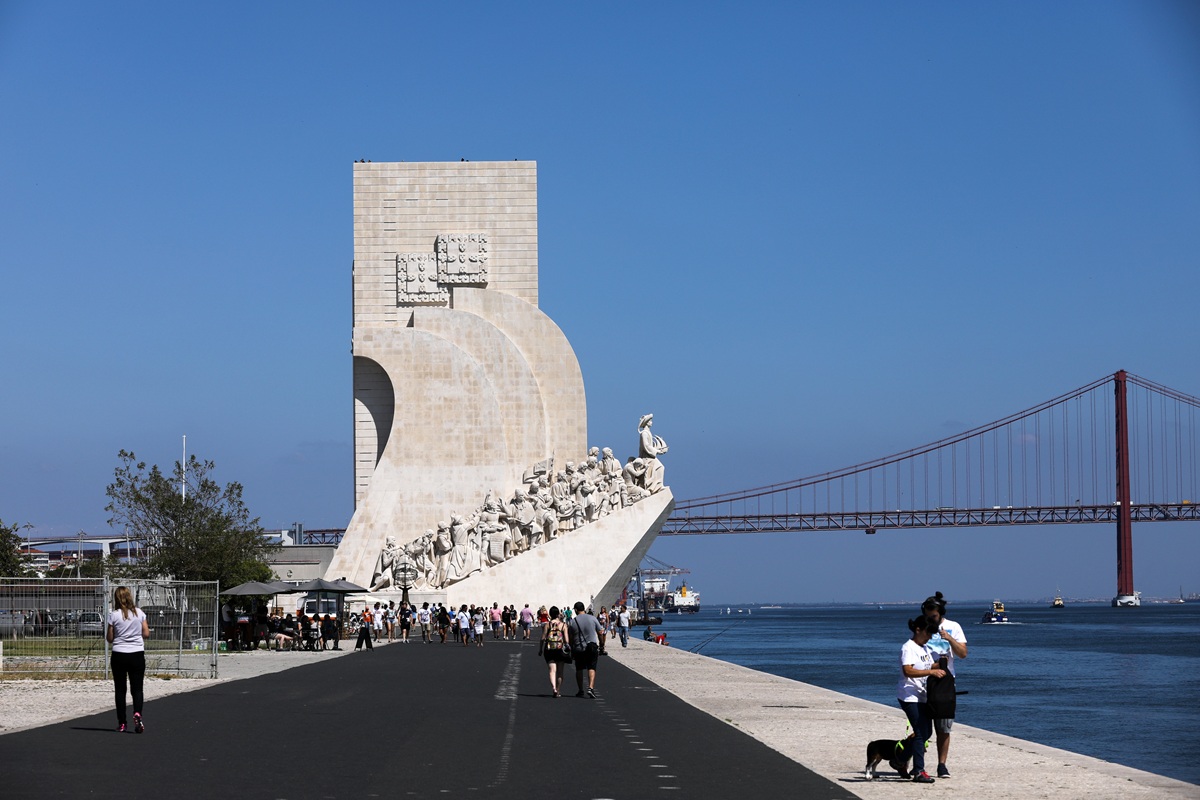 Vista do icónico monumento Padrão dos Descobrimentos ao longo da orla marítima, com pessoas a caminhar e a Ponte 25 de Abril ao fundo.