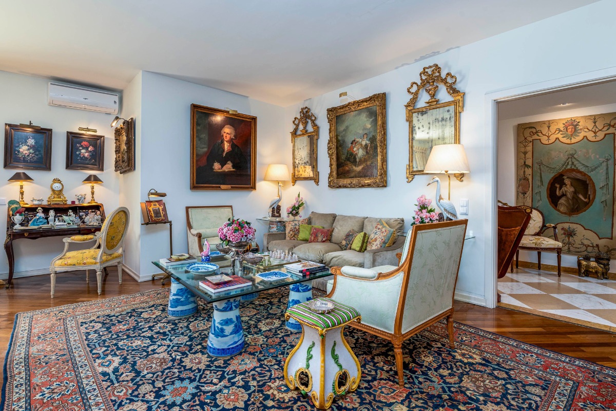 Fotografia de uma sala do Palácio do Correio Velho com mobiliário elegante, pinturas ornamentadas, vasos decorativos e um tapete com padrões ricos. Ambiente acolhedor e convidativo.