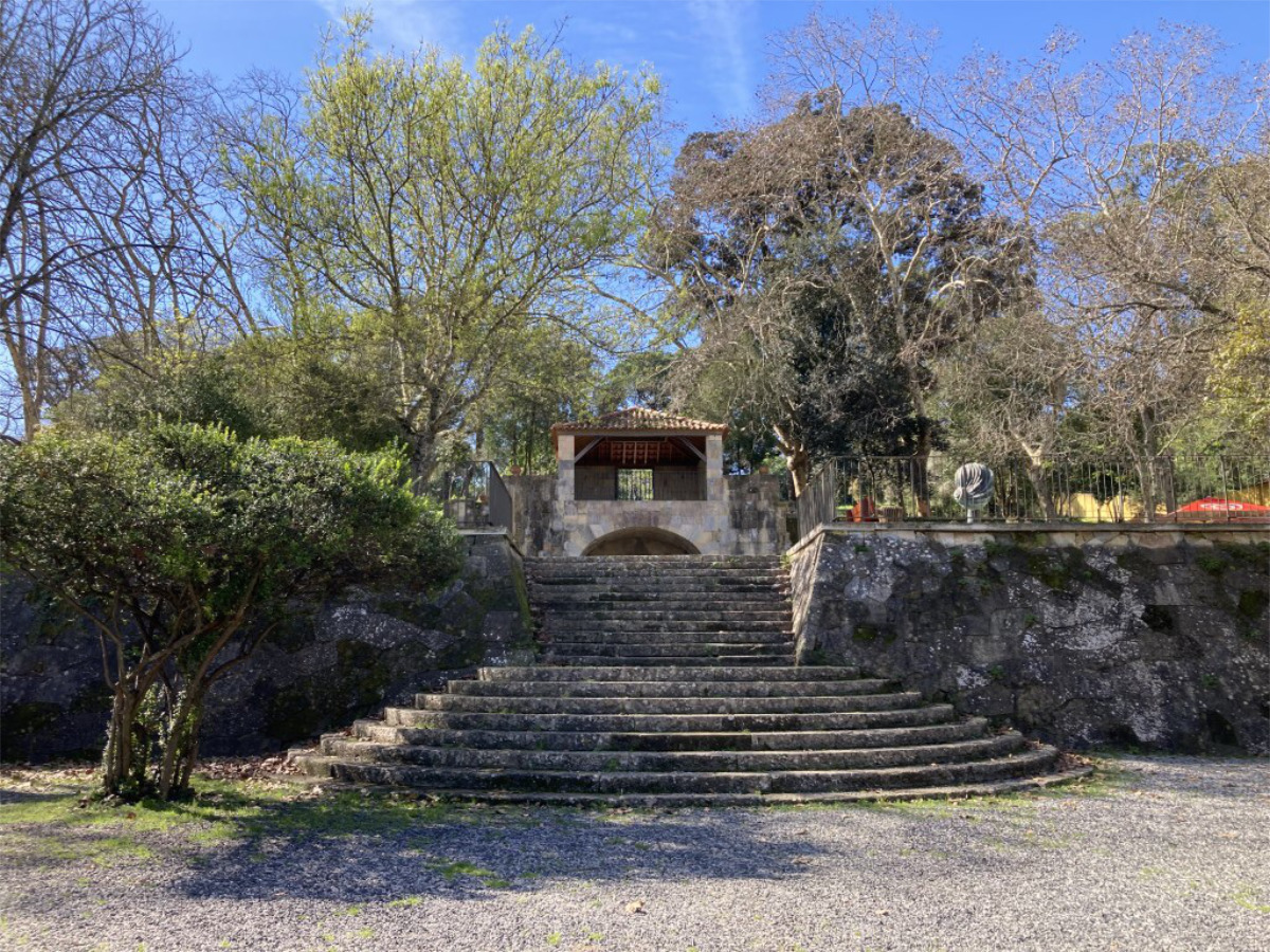 Imagem de uma escadaria antiga de pedra que leva a uma estrutura sob árvores verdes, no Parque Bensaúde, com céu azul ao fundo e um ambiente ao ar livre.