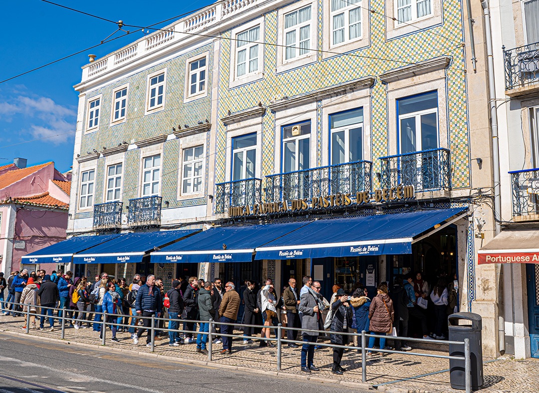 Exterior da famosa pastelaria com uma longa fila de clientes à porta. Edifício revestido a azulejos e com toldos azuis onde se lê "Pastéis de Belém"