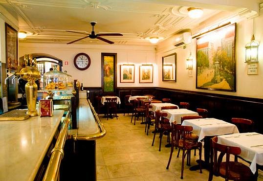 Interior acolhedor da Pastelaria Benard, com decoração vintage, cadeiras e mesas de madeira com toalhas brancas. Iluminação acolhedora, arte nas paredes e um relógio clássico criam uma atmosfera convidativa.